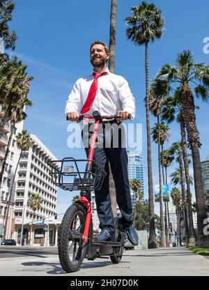 Felice avvocato in formalwear ride e-scooter sul marciapiede, in sella Foto Stock