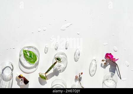 Moisturizer, rullo del viso di giada verde con foglie esotiche di monstera e fiori di orchidea e baubles di vetro. Sole, lunghe ombre. Piatto posare su bianco Foto Stock