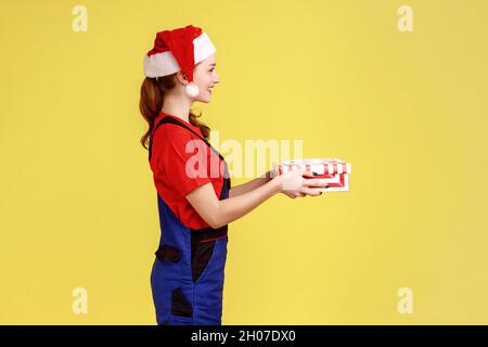 Ritratto della vista laterale di sorridente corriere femmina in piedi che dà la scatola del presente avvolta per il cliente, indossando le tute blu e cappello di babbo natale. Studio interno girato isolato su sfondo giallo. Foto Stock