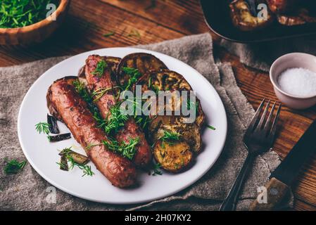 Salsicce di maiale al forno con Eggplant e Leek su piatto bianco Guarnito con erbe fresche Foto Stock