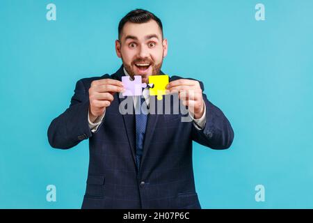 L'uomo bearded eccitato che indossa il vestito ufficiale di stile che tiene e mostra due pezzi del puzzle, matching e collegando puzzle, risolvere problemi, creatività. Studio interno girato isolato su sfondo blu. Foto Stock