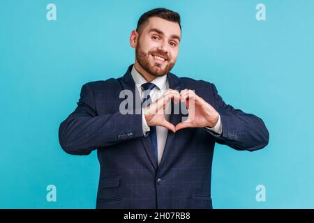 Felice bell'uomo portico indossando un abito ufficiale in piedi con cuore o gesto d'amore e guardando la macchina fotografica con un sorriso toothy. Studio interno girato isolato su sfondo blu. Foto Stock