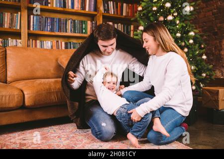 Tema delle vacanze di Natale e Capodanno in famiglia con bambino piccolo. Famiglia di tre, avvolta in coperta, si siederà sul pavimento nel soggiorno vicino Foto Stock