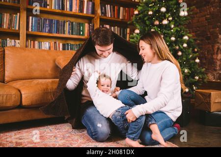 Magica atmosfera di natale in casa accogliente. La giovane famiglia con bambino ama giocare sul piano coperto di coperta vicino all'albero di Natale durante le vacanze di Capodanno Foto Stock