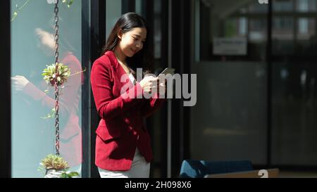 Donna d'affari in tuta rossa appoggiata su una finestra di vetro durante la lettura o il messaggio di SMS su smartphone cellulare in ufficio Foto Stock