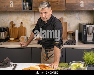 Ritratto uno chef elegante in una giacca nera posa in cucina. Lo chef allegro di etnia caucasica prepara il cibo nella sua cucina. Persone al lavoro. Pro Foto Stock