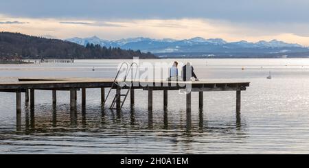 Herrsching, Germania - 22 gennaio 2021: Due persone sedute su un molo di legno sul lago Ammersee. Alpi sullo sfondo. Foto Stock