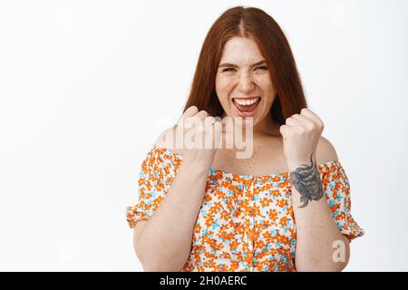 La ragazza rossa entusiasta urla e stringe i pugni in gioia, vincendo o conseguendo l'obiettivo, celebrando la vittoria, in piedi in abito su sfondo bianco Foto Stock