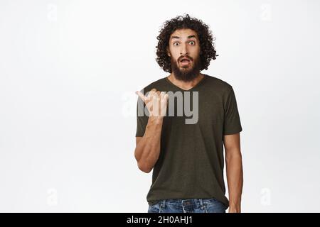 Sorpreso giovane uomo che punta a sinistra, gasping e fissando scioccato alla macchina fotografica, mostrando grande promozione, pubblicità, in piedi su sfondo bianco Foto Stock