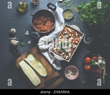 Lasagne di zucchine fatte in casa con basilico fresco, bolognese, pomodori, aglio, olio d'oliva e formaggio in piatto da forno bianco su tavolo da cucina in cemento scuro. Co Foto Stock