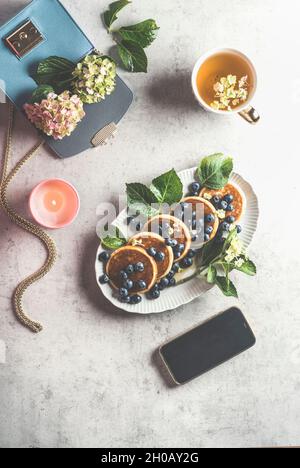 Stile di vita femminile per la colazione con pancake mirtilli, tazza di tè, candela e smartphone con schermo nero, borsa blu con hydrangea su l Foto Stock