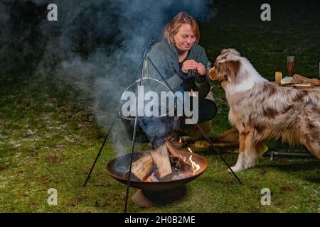 Giovane donna bionda sorridente dà biscotti al suo cane tricolore Australian Shepherd. In inverno, accanto al bollitore per fumare sul fuoco, al buio Foto Stock