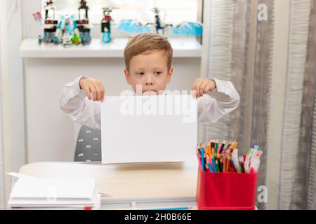 Un ragazzo carino prima livellatore in una divisa della scuola a casa mentre isolato alla sua scrivania tiene un foglio di carta bianca. Messa a fuoco selettiva. Verticale Foto Stock