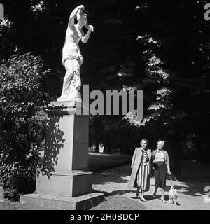 Zwei junge Frauen mit Foxterrier kommen un einer statua in einem Park a Stoccarda vorbei, Deutschland 1930er Jahre. Due giovani donne e un fox terrier passeggiando in un giardino pubblico a Stoccarda, Germania 1930s. Foto Stock