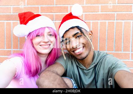 buon natale! felice coppia variegata di amici che indossano natale santa cappello facendo un ritratto selfie pov seduta su una parete di fondo insieme Foto Stock