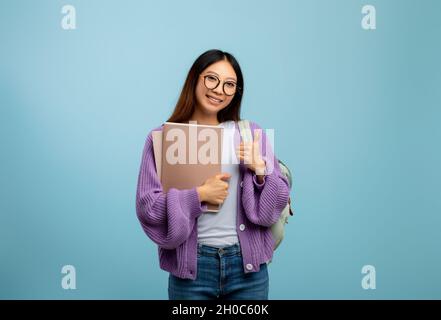 Positiva studentessa asiatica che tiene libri di lavoro e mostra il pollice in su, sorridendo alla macchina fotografica su sfondo blu Foto Stock