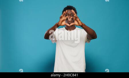 Uomo gioioso che fa il simbolo della forma del cuore con le mani mentre guarda la macchina fotografica. Persona romantica che mostra segno d'amore per giorno di San Valentino. Adulto che celebra il romanticismo con un gesto affettuoso. Foto Stock