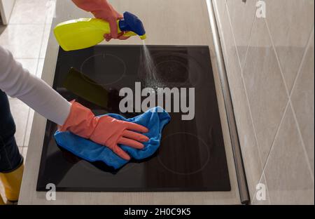 Primo piano delle mani femminili con guanti protettivi spruzzando stufa a induzione e pulendo con panno in cucina Foto Stock