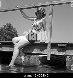 Eine junge Frau im Badeanzug sitzt auf einem Holzsteg un einem vedere, Deutschland 1930er Jahre. Una giovane donna che indossa il costume da bagno siiting su una passerella sulla riva di un lago, Germania 1930s. Foto Stock