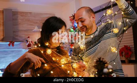 Uomo e donna aggrovigliati in ghirlanda di luci scintillanti mentre decorano per la festa della vigilia di natale. Coppia ridendo e untangling nodo di archi luci con lampadine illuminate. Gente allegra Foto Stock