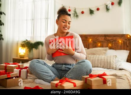 Felice afroamericana signora che festeggia l'anno nuovo seduto a letto e abbracciando la scheda di Natale con gli occhi chiusi Foto Stock