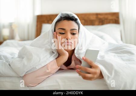 Giovane donna nera sconvolta che guarda lo schermo dello smartphone mentre si stesa nel letto sotto la coperta, sentendosi solitaria o depressa Foto Stock