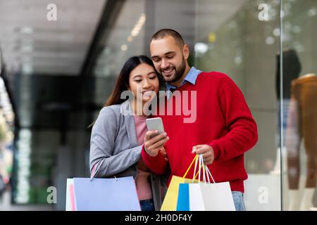 Allegro giovane coppia interrazziale con borse regalo e cellulare ordinare merci online vicino centro commerciale Foto Stock