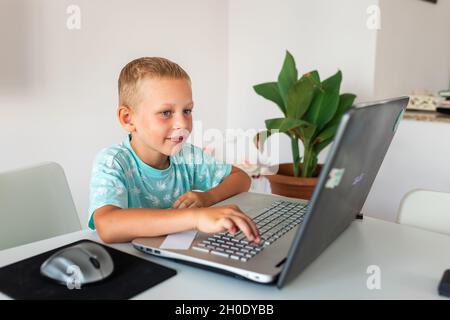 Piccolo ragazzo di scuola giovane che lavora a casa con un laptop e appunti di classe che studiano in una classe virtuale che ha una videoconferenza su laptop con l'insegnante da Foto Stock