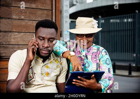 una coppia turistica africana che si gode tour ed esplorare la città l'uomo al telefono la donna guardare lo schermo del suo tablet tutti felice ben vestito Foto Stock