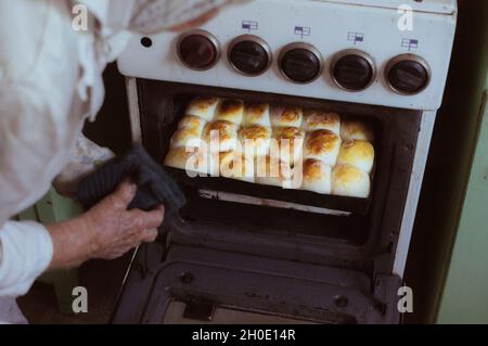 Una donna di 90 anni toglie dal forno una teglia da forno con torte ruddy cotte Foto Stock