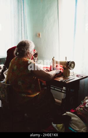 Una vecchia donna groppa dai capelli grigi con capelli ricci cucita su una macchina da cucire retrò d'epoca Foto Stock