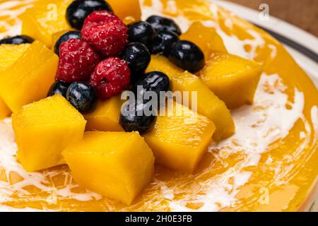 Alto angolo di vista closeup di mango cheesecake guarnito con mucchio di frutta biologica, pezzi di mango mature, lampone conservato, mirtillo e grappa matura Foto Stock