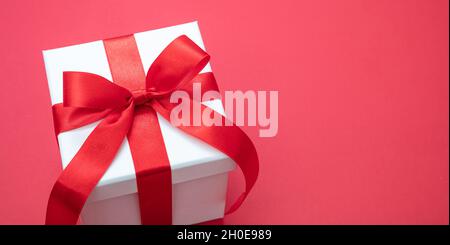 Scatola regalo bianco Natale arco legato con nastro su sfondo rosso, sorpresa di San Valentino, regalo di Capodanno, satin ricci decorazione. Copia spac Foto Stock