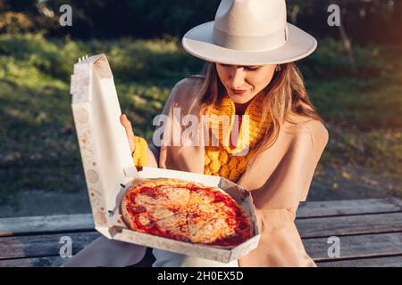 Ritratto di giovane donna alla moda che ha la pizza nel parco autunnale. Scatola apribile con pepperoni all'aperto. Mangiare uno spuntino caldo Foto Stock