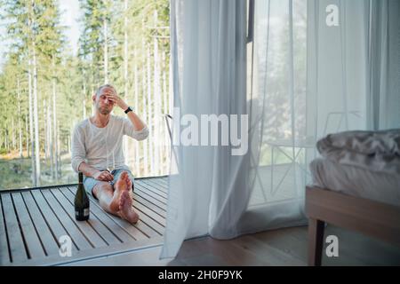 Ritratto di un uomo stanco di mezza età seduto a piedi nudi sul pavimento della casa forestale balcone, egli bere vino bianco e massaggiando templi. Persone esauste o m Foto Stock
