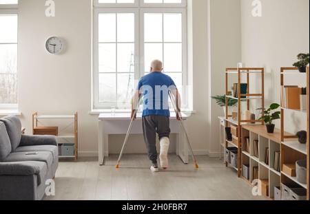 Uomo anziano con gamba rotta in gesso calco utilizzando stampelle e camminare da solo a casa Foto Stock