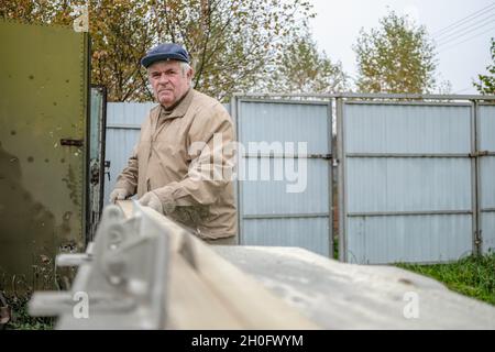 Un vecchio carpentiere dai capelli grigi taglia un asse su una macchina con una sega circolare. Messa a fuoco selettiva. Un disco rotante di sega sega un pannello di legno lungo. Un uomo anziano professionista lavora all'aperto. Foto Stock