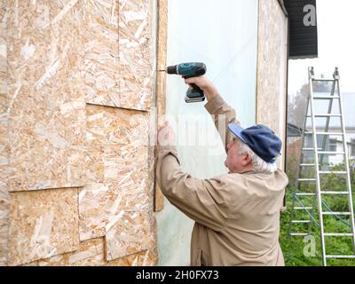 Un uomo anziano dai capelli grigi avvita un asse orientato del trefolo alla parete. Il carpentiere stringe le viti con un cacciavite a batteria. Costruzione di una casa di legno di campagna. Foto Stock