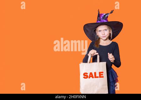 Una bella ragazza sorridente in un abito nero e un cappello strega tiene una borsa di carta artigianale con le parole VENDITA su uno sfondo arancione. Shopping di Halloween conc Foto Stock