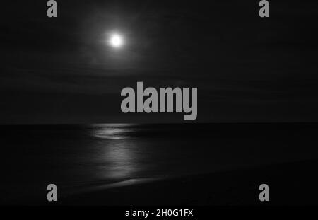 Notte di mare e surf in bianco e nero con la luna luminosa che brilla sulla calme surf e la costa. Foto Stock