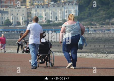 PERSONA IN SOVRAPPESO CHE CAMMINA SUL LUNGOMARE DI LLANDUDNO GALLES REGNO UNITO RE OBESITÀ ESERCIZIO CIBO ECC Foto Stock