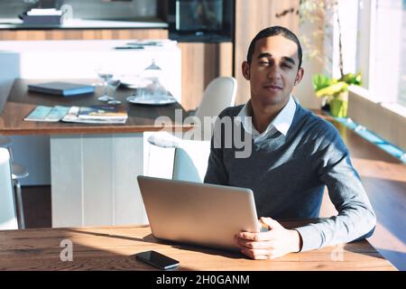 Giovane uomo nero che sta fissando la fotocamera con un sorriso mentre si siede a un tavolo che lavora su un computer portatile in un raggio di luce solare Foto Stock