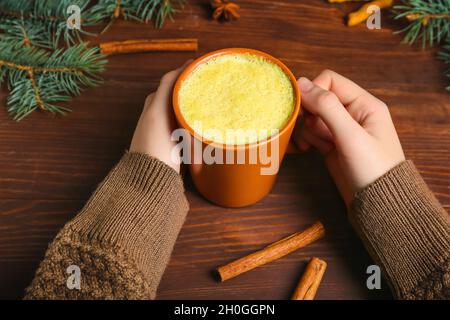 Mani femminili con tazza di gustoso latte di curcuma, rami di cannella e abete su sfondo ligneo Foto Stock