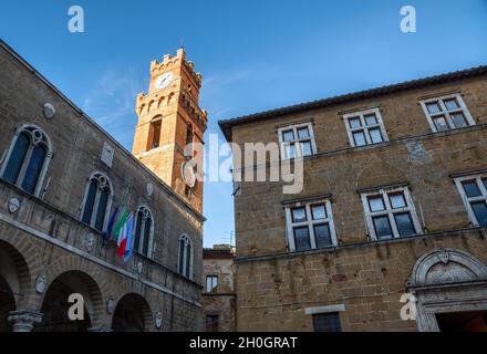 Pienza, Toscana, Italia. Agosto 2020. Il sole del tardo pomeriggio illumina la torre dell'orologio nel centro storico con una luce calda. Sotto le bandiere sul Foto Stock