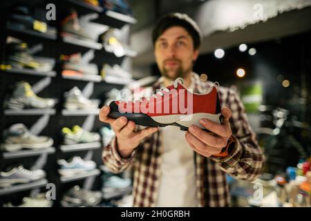 Consulente del venditore maschile caucasico in negozio all'aperto che presenta scarpe da trekking al compratore. Un venditore in un negozio di articoli sportivi aiuta un acquirente a scegliere le escursioni Foto Stock