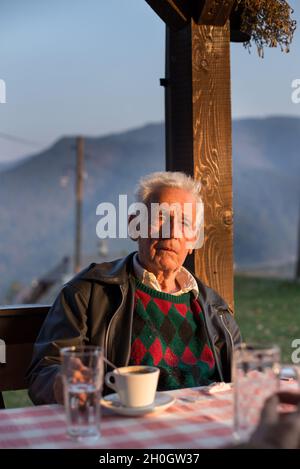 Ritratto di vecchio uomo seduto in un caffè con montagne sullo sfondo Foto Stock