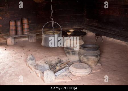 Vecchio camino in cottage di legno con calderone di ferro appeso sopra e vasi di argilla intorno Foto Stock