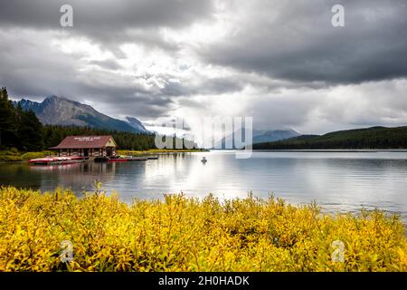 Curly Phillips Boathouse, storica boathouse sulla riva del lago Maligne, dietro di esso catena montuosa Queen Elizabeth Ranges, vegetazione autunnale, nuvoloso Foto Stock