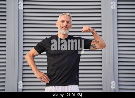 Uomo di mezza età sicuro che guarda la macchina fotografica, mostrando il suo bicipite mentre si posa all'aperto su sfondo grigio Foto Stock