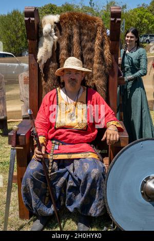 Eungella, Queensland, Australia - Ottobre 2021: Uomo in replica costume vichingo seduta in una sedia in legno fur foderato con cameriera medievale dietro di lui a re- Foto Stock